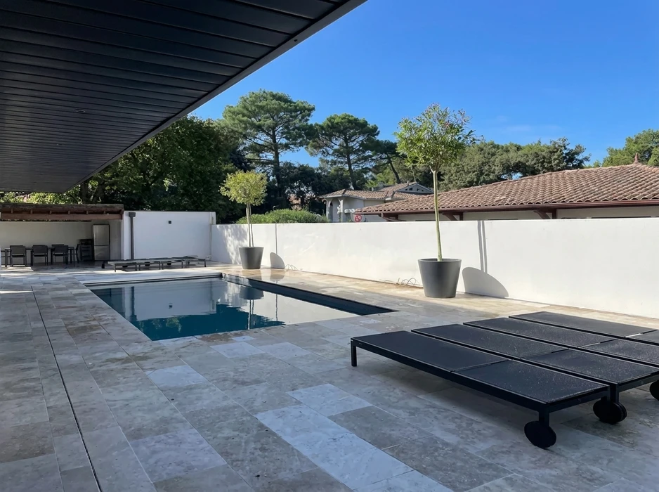 Piscine extérieure rectangulaire à débordement partiel, dallage en travertin beige, transats noirs à roulettes design contemporain